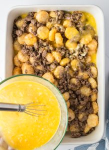 A casserole dish filled with cooked ground beef and tater tots, with a bowl of beaten eggs being poured over the top.