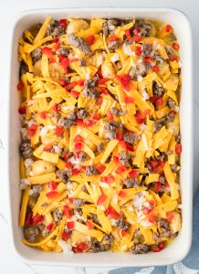 Casserole with shredded cheese, ground meat, red peppers, onions, and potato pieces in a white baking dish.