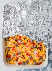 A casserole dish filled with cheese, ground meat, diced tomatoes, and onions, partially covered with aluminum foil.