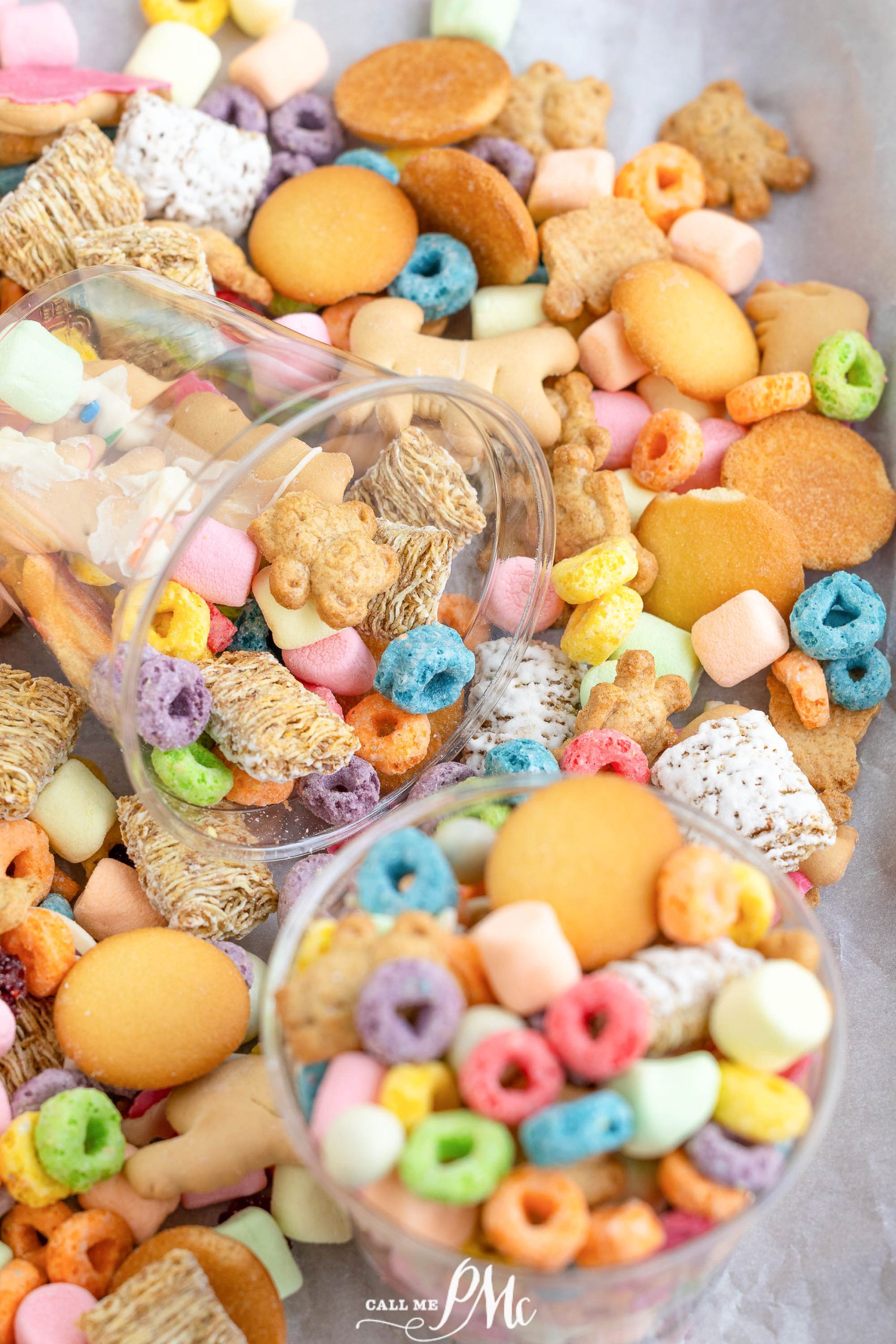 Two clear containers filled with colorful cereal and snacks, including marshmallows, cookies, and cereal rings, spilled onto a surface with a mix of textures and colors spread around.