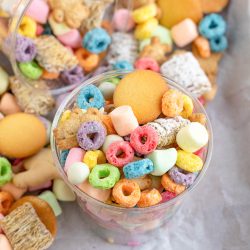 A clear cup filled with assorted colorful cereals, marshmallows, animal crackers, and wafers, placed on a parchment-lined surface with more snacks scattered around.