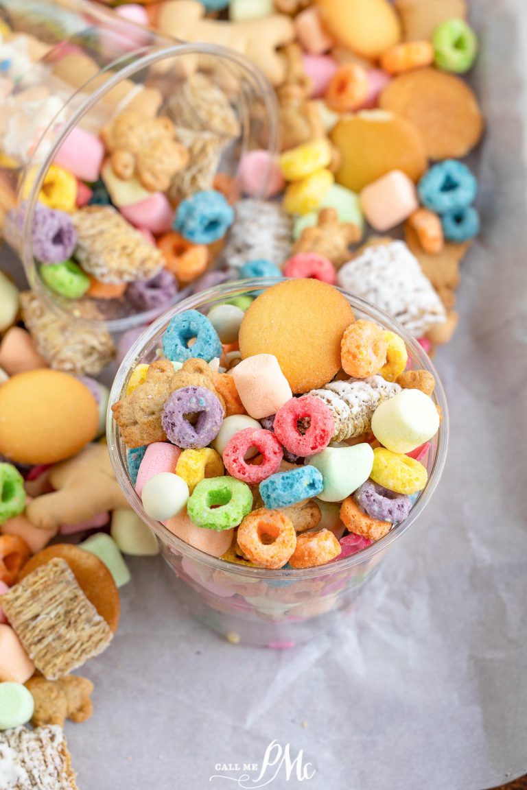 A clear cup filled with assorted colorful cereals, marshmallows, animal crackers, and wafers, placed on a parchment-lined surface with more snacks scattered around.