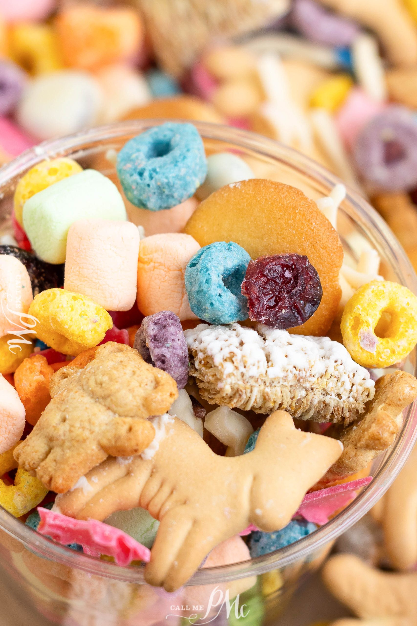 Close-up of a snack mix with colorful cereal loops, marshmallows, animal crackers, a dried cranberry, and other assorted treats in a clear bowl.