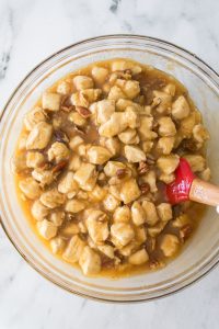 Glass bowl filled with dough pieces coated in a caramel sauce with pecans, stirred with a red silicone spatula.