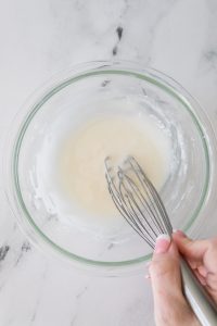 A hand whisking a white mixture in a clear glass bowl on a marble surface.