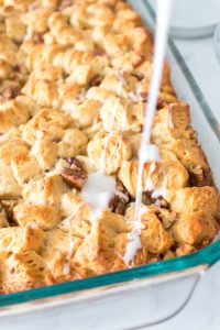 A freshly baked bread pudding in a glass dish, topped with pecans, is being drizzled with white glaze.