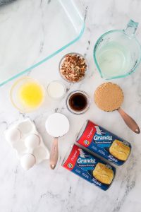 Ingredients laid out on a marble countertop, including eggs, canned biscuits, brown sugar, butter, pecans, vanilla, flour, and milk, next to a glass baking dish.