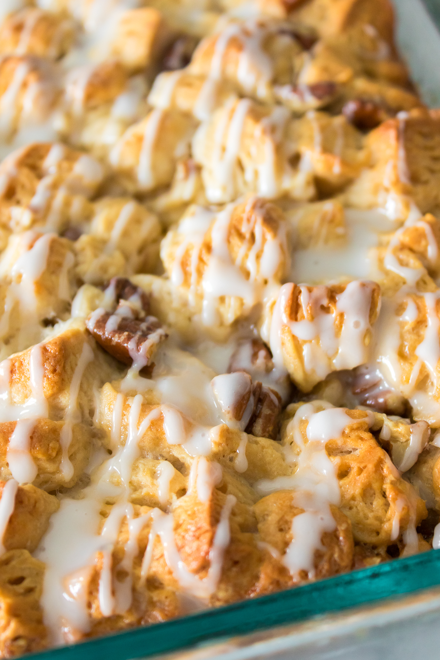 Close-up of a baked pastry dessert with a golden-brown surface, drizzled with white icing and pieces of chocolate throughout, in a glass baking dish.