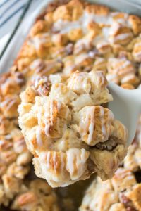 Close-up of a glazed monkey bread piece being lifted from a tray with a white spatula. The bread appears golden brown and topped with icing.