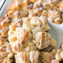 Close-up of a glazed monkey bread piece being lifted from a tray with a white spatula. The bread appears golden brown and topped with icing.