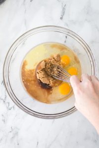 Hand whisking eggs, brown sugar, and liquid ingredients in a glass bowl on a marble countertop.