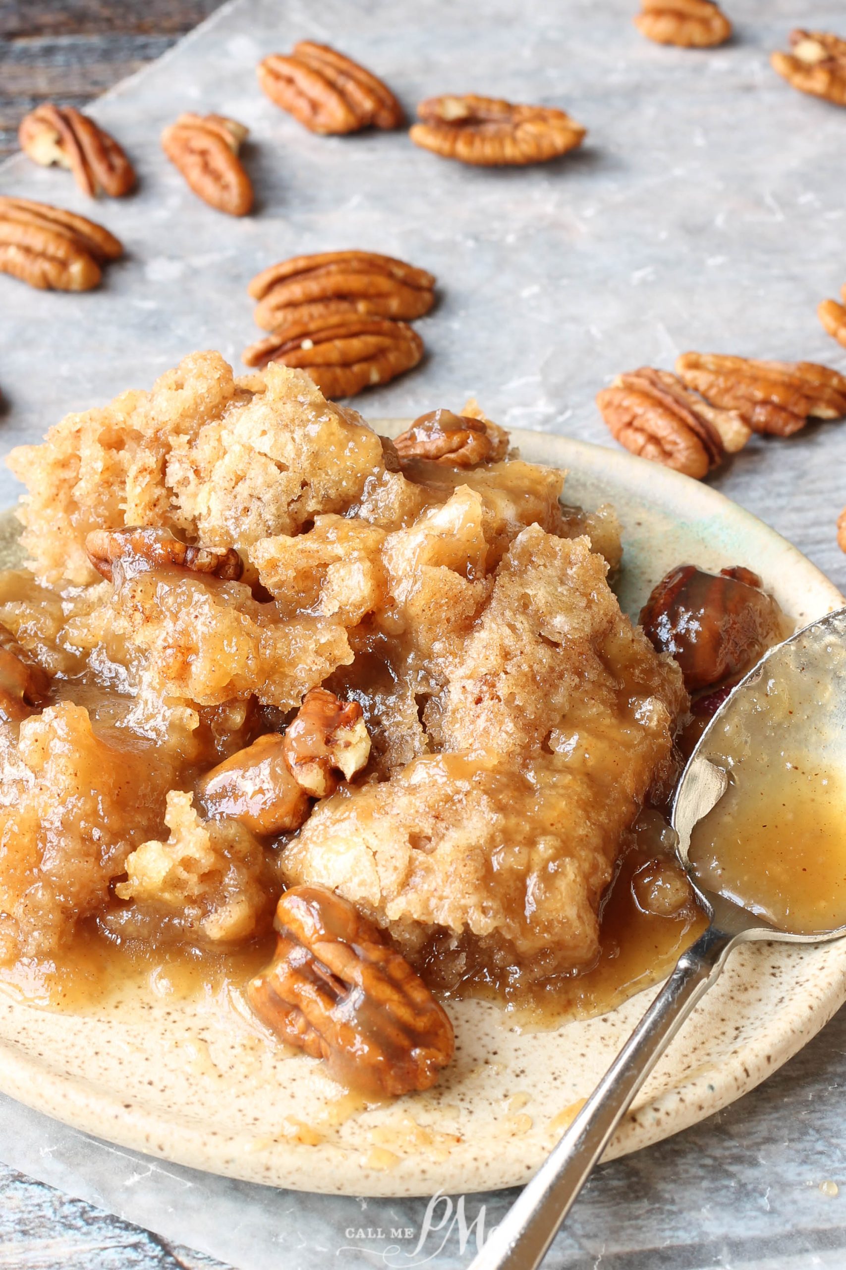 A plate of pecan cobbler drizzled with brown butter sauce, topped with pecan halves. A spoon rests beside the cobbler, while several pecans are scattered on a parchment-lined surface.