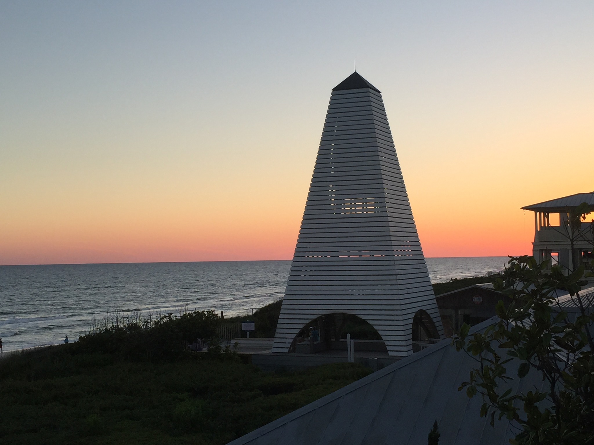 Sunset over a beach with a tall, striped, pyramid-like structure in the foreground and a building on the right.