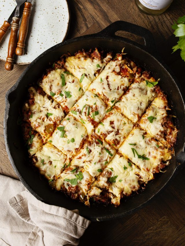 A cast iron skillet filled with cut squares of cheesy lasagna topped with parsley, next to empty plates and utensils.