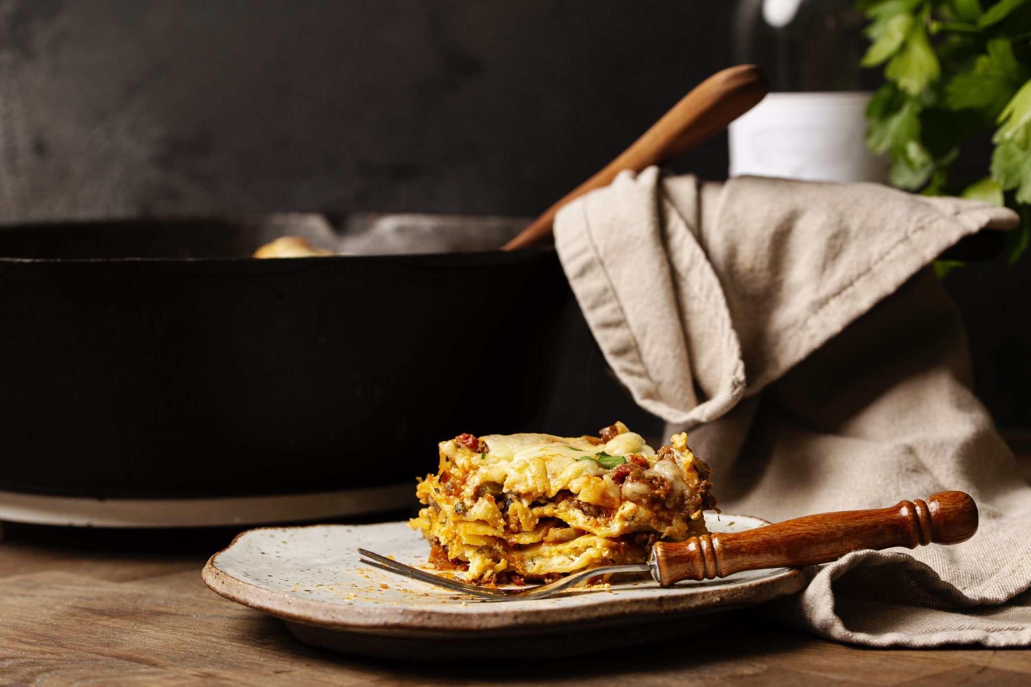 A slice of lasagna on a plate with a fork, near a pan and a cloth. A bottle and parsley are in the background.