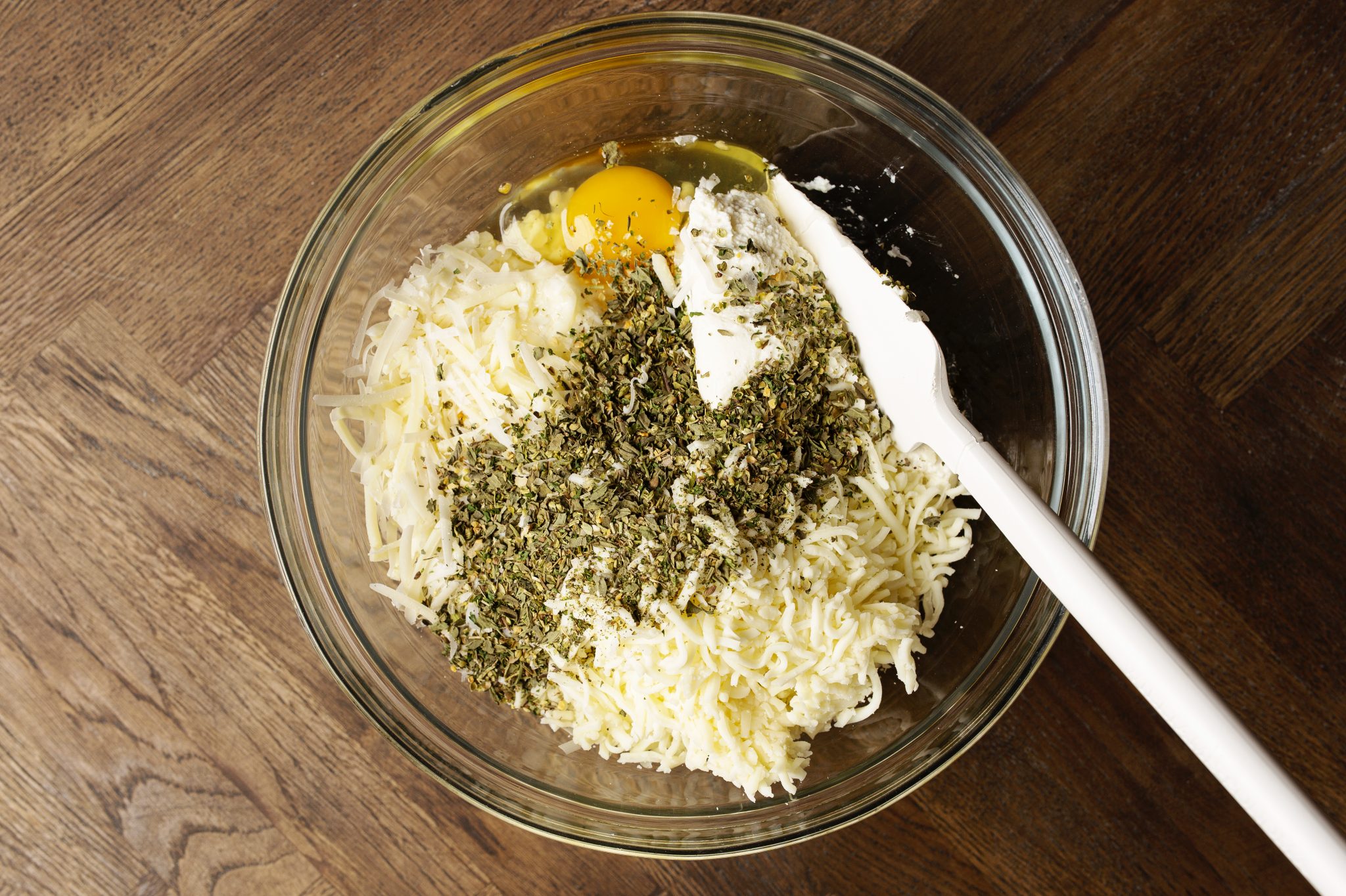 A clear glass bowl on a wooden surface contains shredded cheese, an egg, and herbs, with a white spatula resting inside.