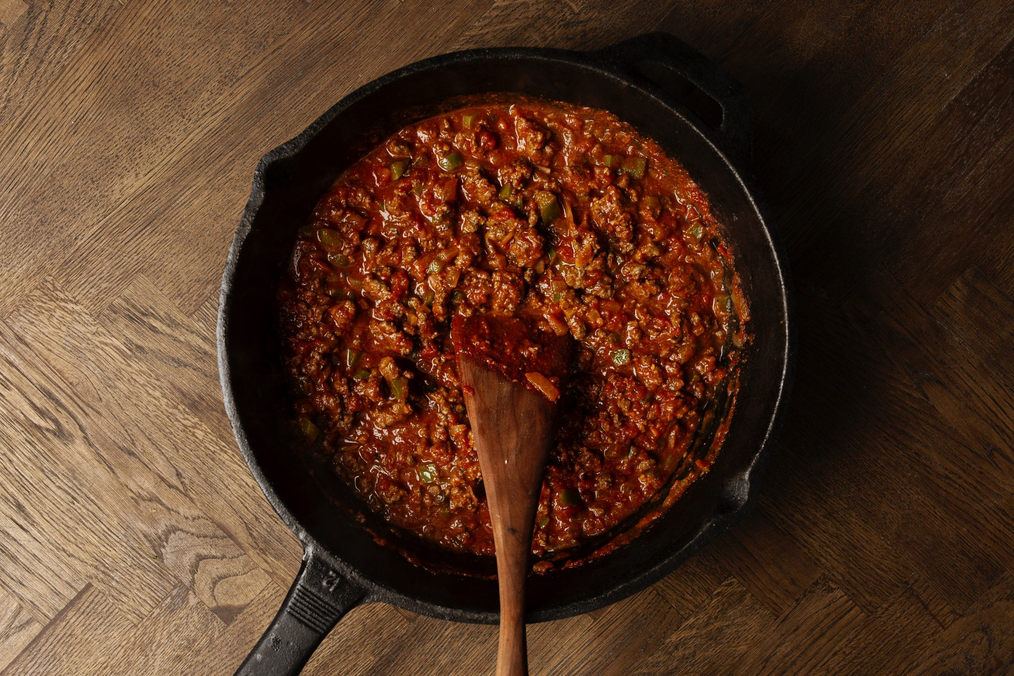 A cast iron pan filled with a chunky tomato-based sauce, mixed with vegetables and ground meat, placed on a wooden surface. A wooden spatula rests in the pan.