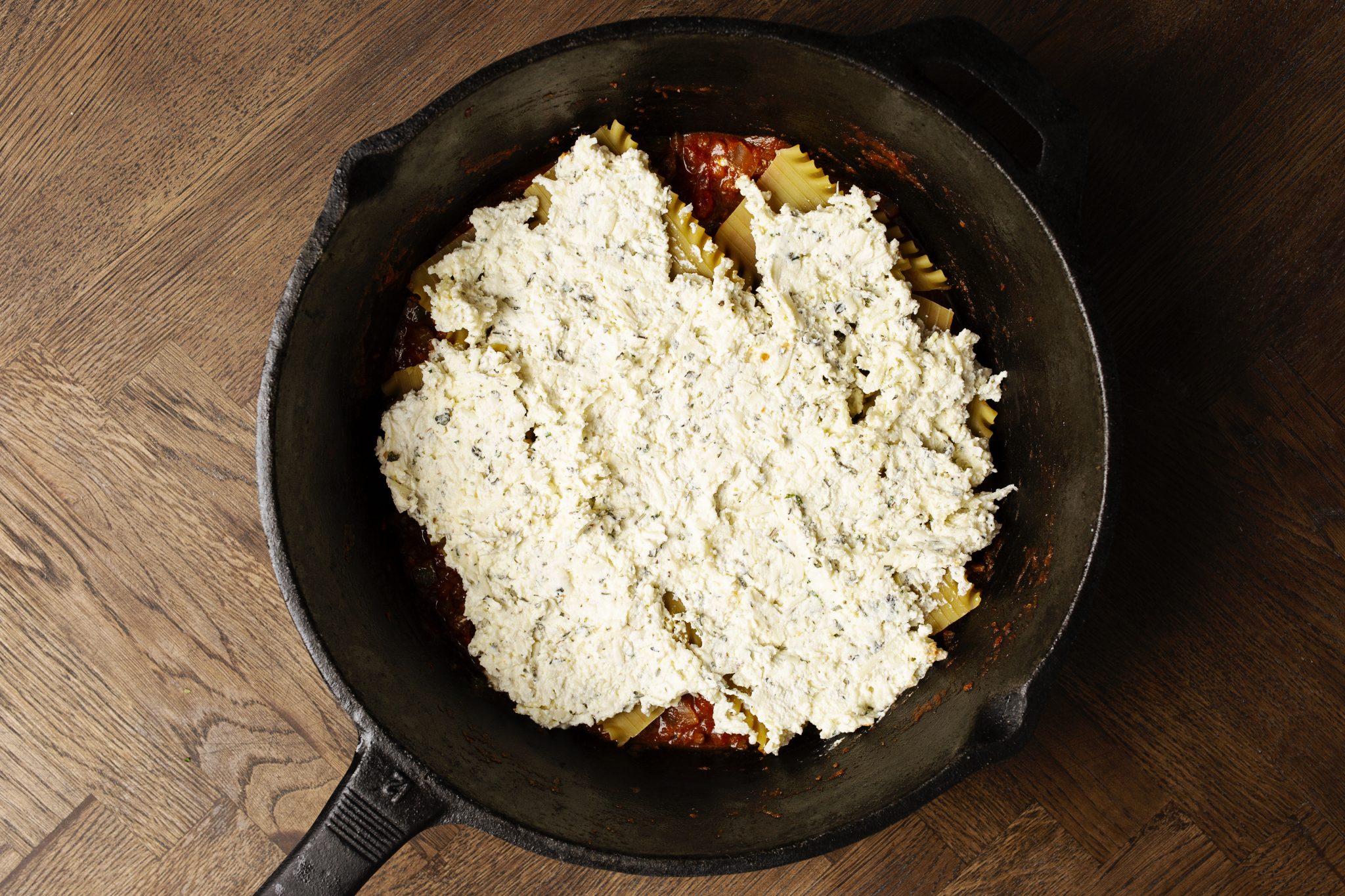 A cast iron pan filled with lasagna ingredients, showing layers of pasta, tomato sauce, and ricotta cheese, placed on a wooden surface.