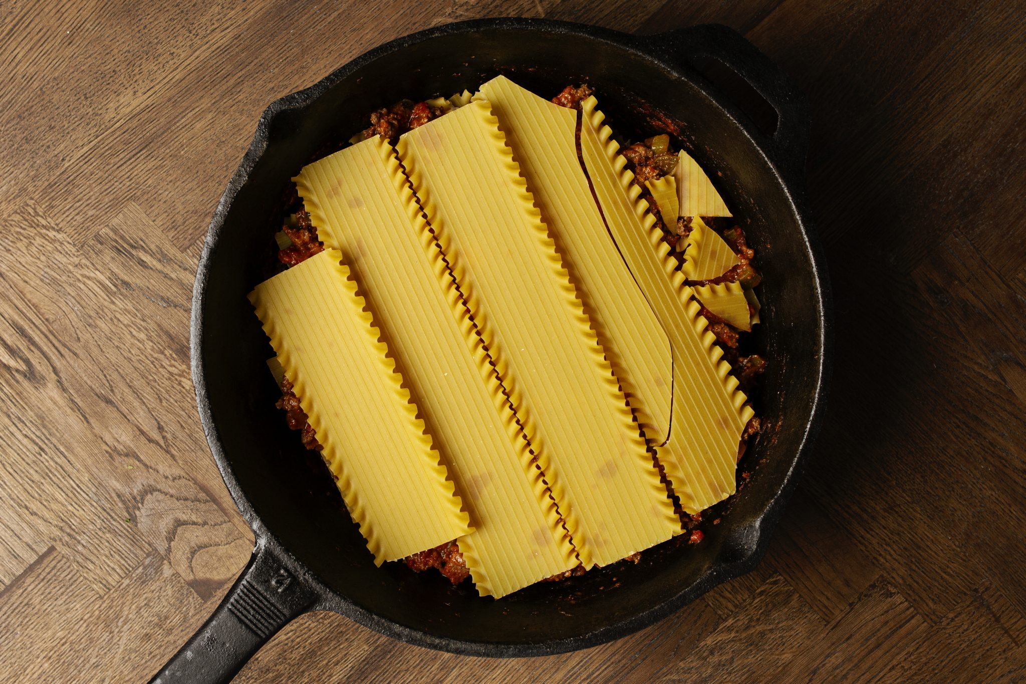 Uncooked lasagna noodles layered over meat sauce in a black skillet on a wooden surface.