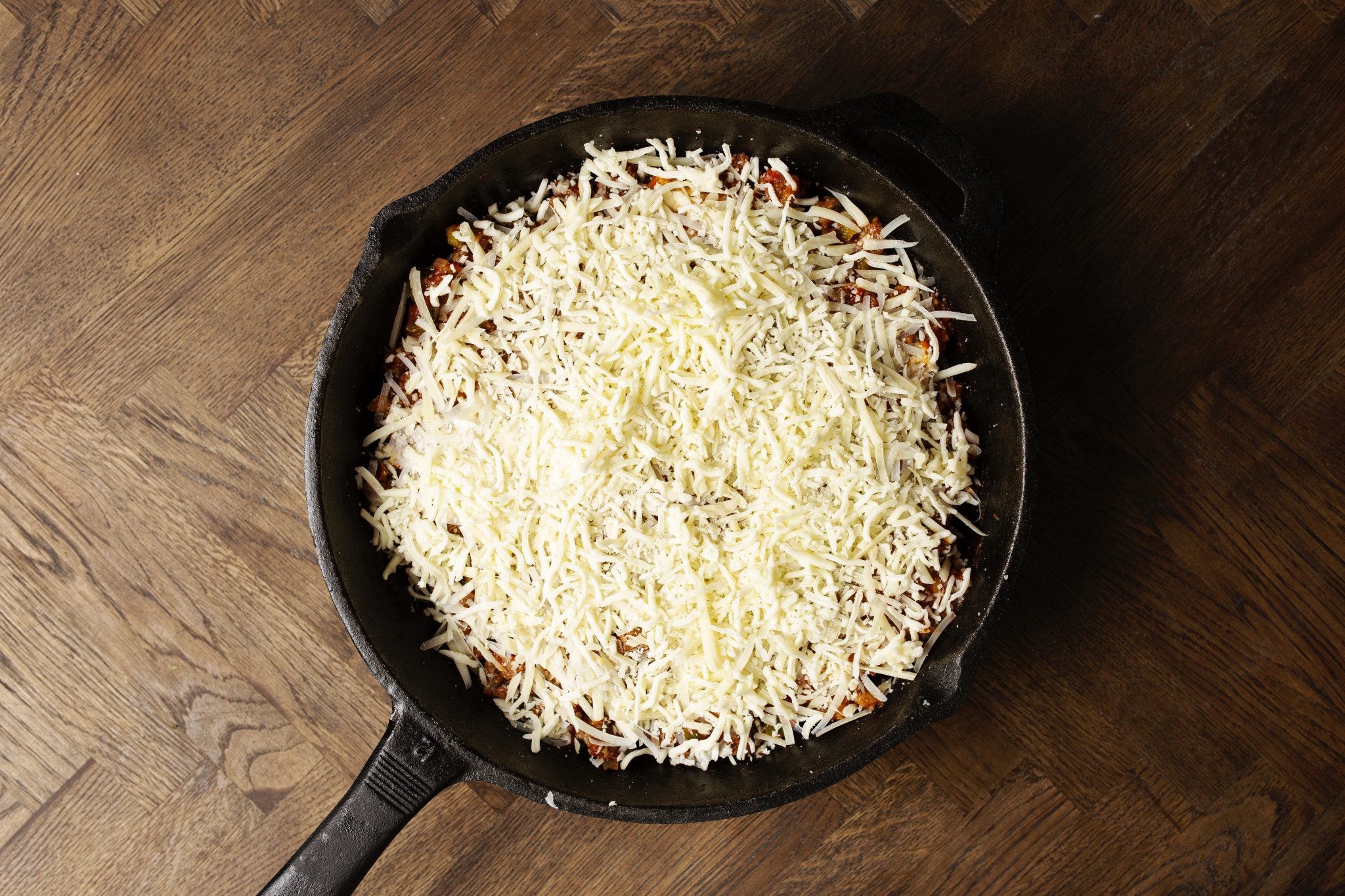 A cast iron skillet filled with a dish topped with shredded cheese on a wooden surface.