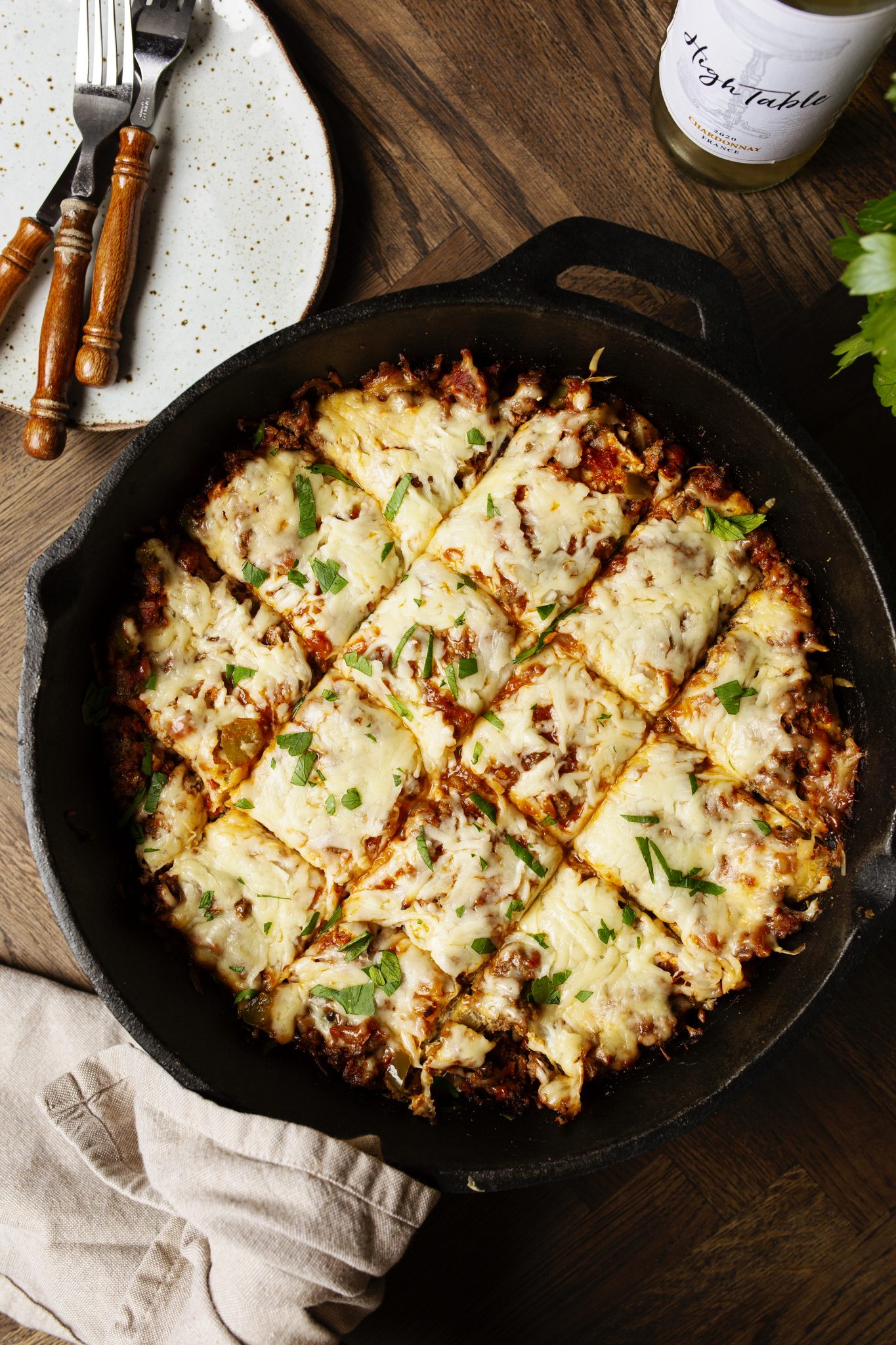 A skillet filled with sliced cheesy casserole topped with herbs, next to a wooden-handled fork and knife, a plate, a napkin, and a jar.