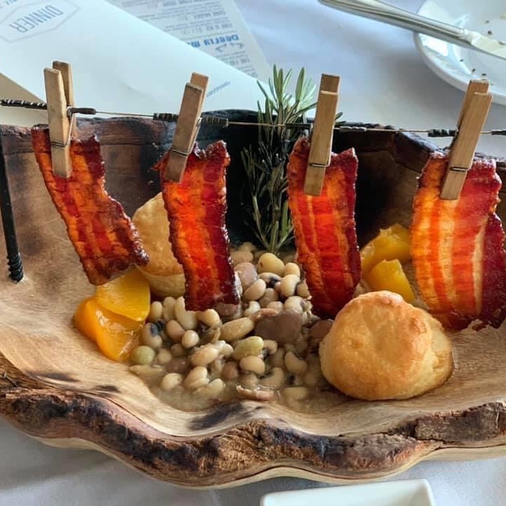 Bacon strips are clipped to a rope above a wooden bowl containing beans, peaches, and bread rolls.