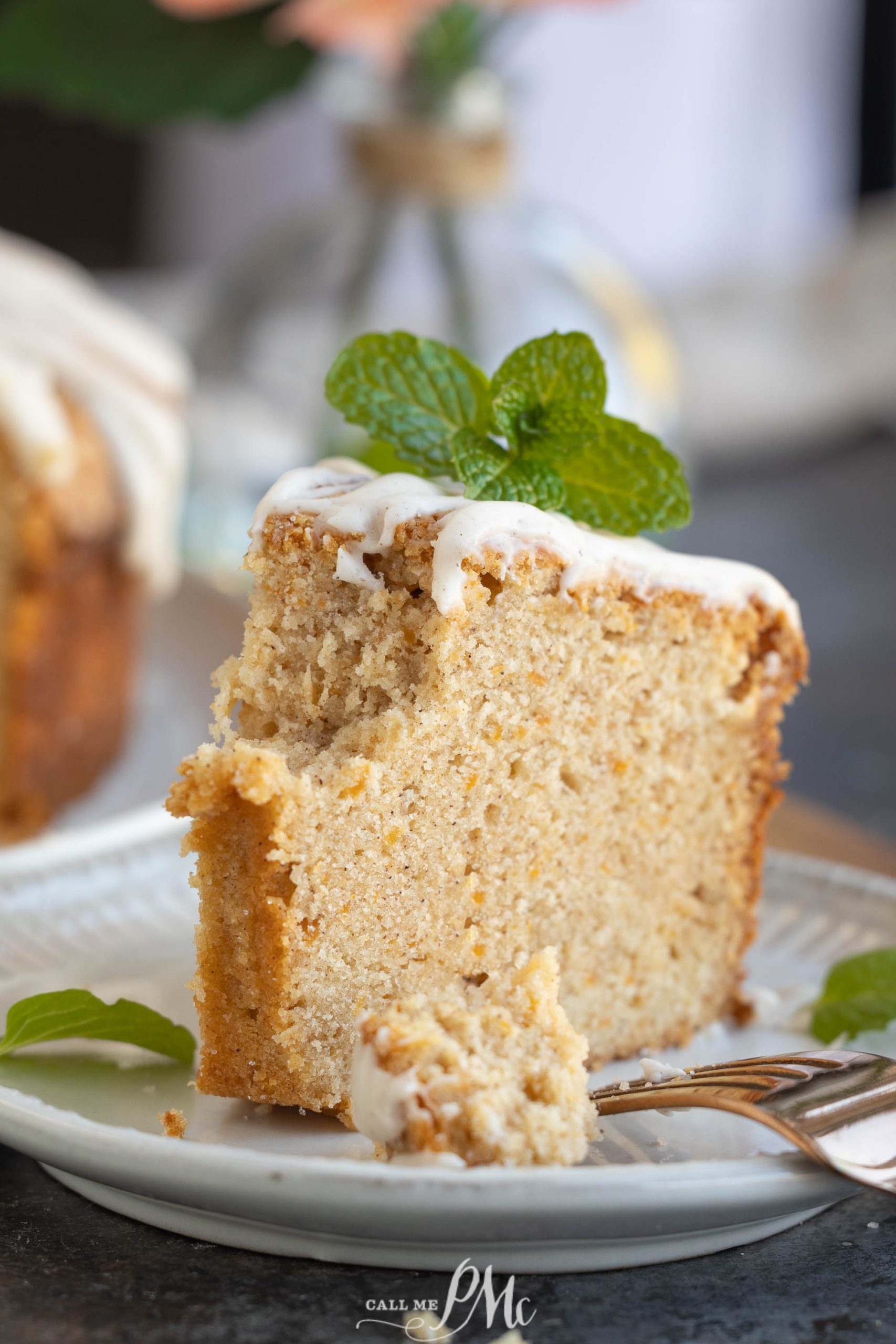 A slice of sweet potato pound cake with white icing and a mint garnish on a white plate, accompanied by a fork holding a delectable piece in the foreground.