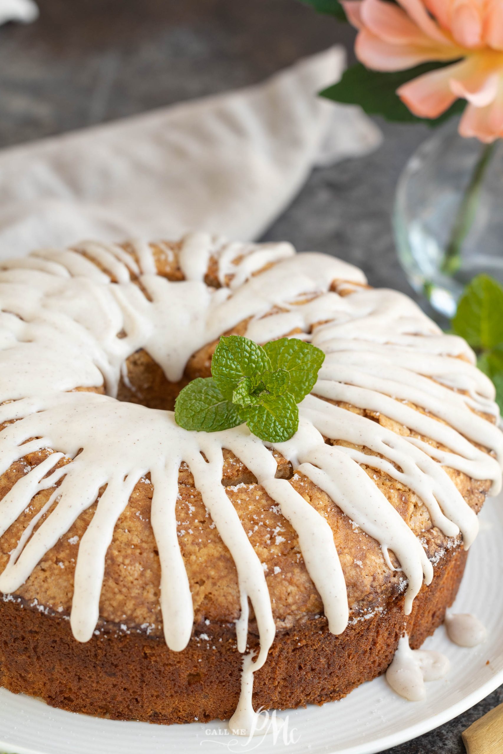 A Sweet Potato Pound Cake is adorned with a drizzle of white icing and fresh mint leaves, elegantly displayed on a white plate. A pink flower and cloth add charm to the background.