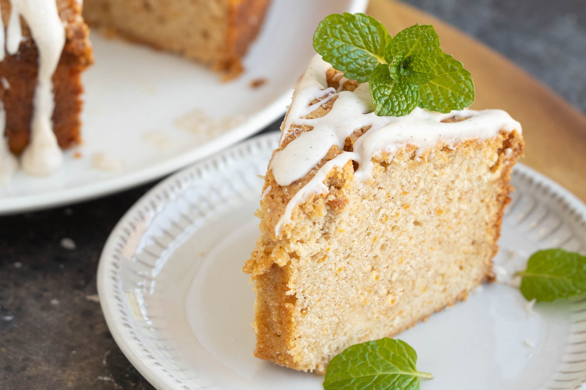 A slice of sweet potato pound cake topped with white icing and a sprig of mint sits elegantly on a white plate. Another slice is tempting in the background.