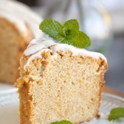 A slice of sweet potato pound cake rests on a white plate, topped with white icing and garnished with mint leaves. A glass bottle is blurred in the background, adding a touch of mystery to this delightful scene.