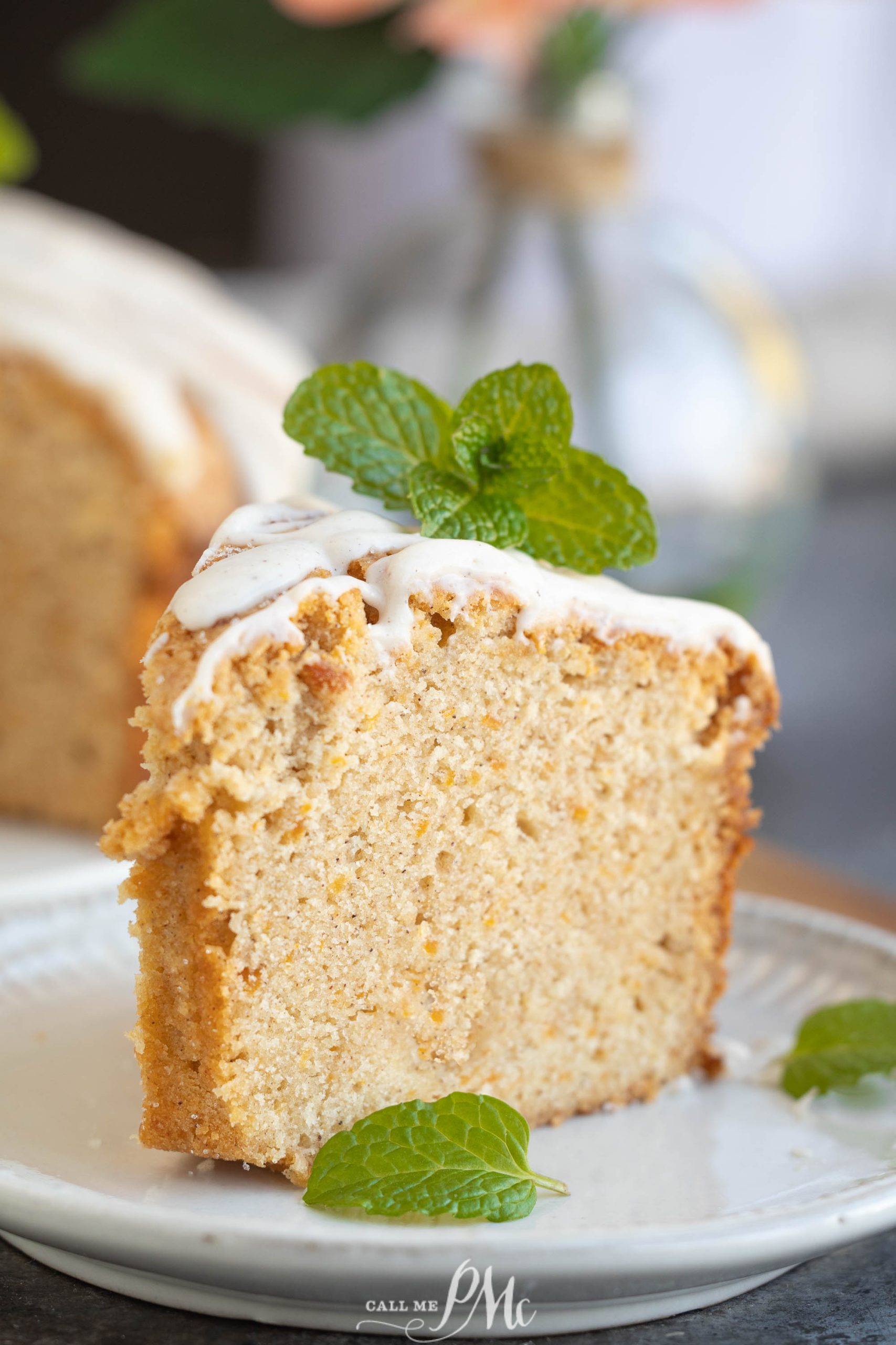 A slice of sweet potato pound cake rests on a white plate, topped with white icing and garnished with mint leaves. A glass bottle is blurred in the background, adding a touch of mystery to this delightful scene.