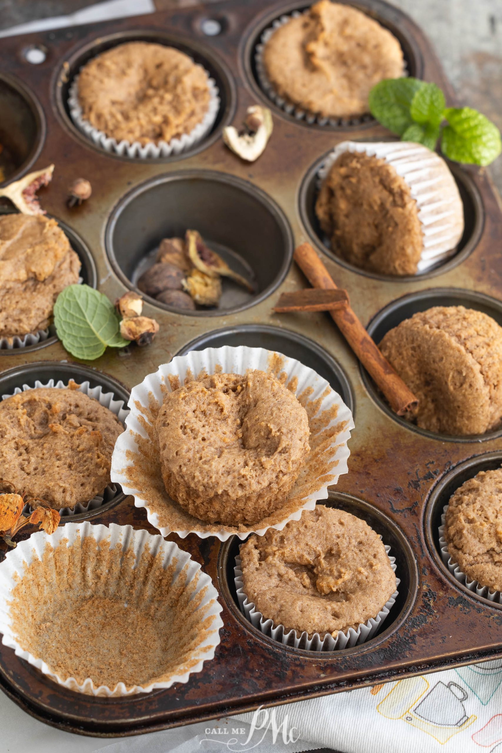 Muffin tin with freshly baked muffins and a few unwrapped. Cinnamon sticks and mint leaves are scattered around.