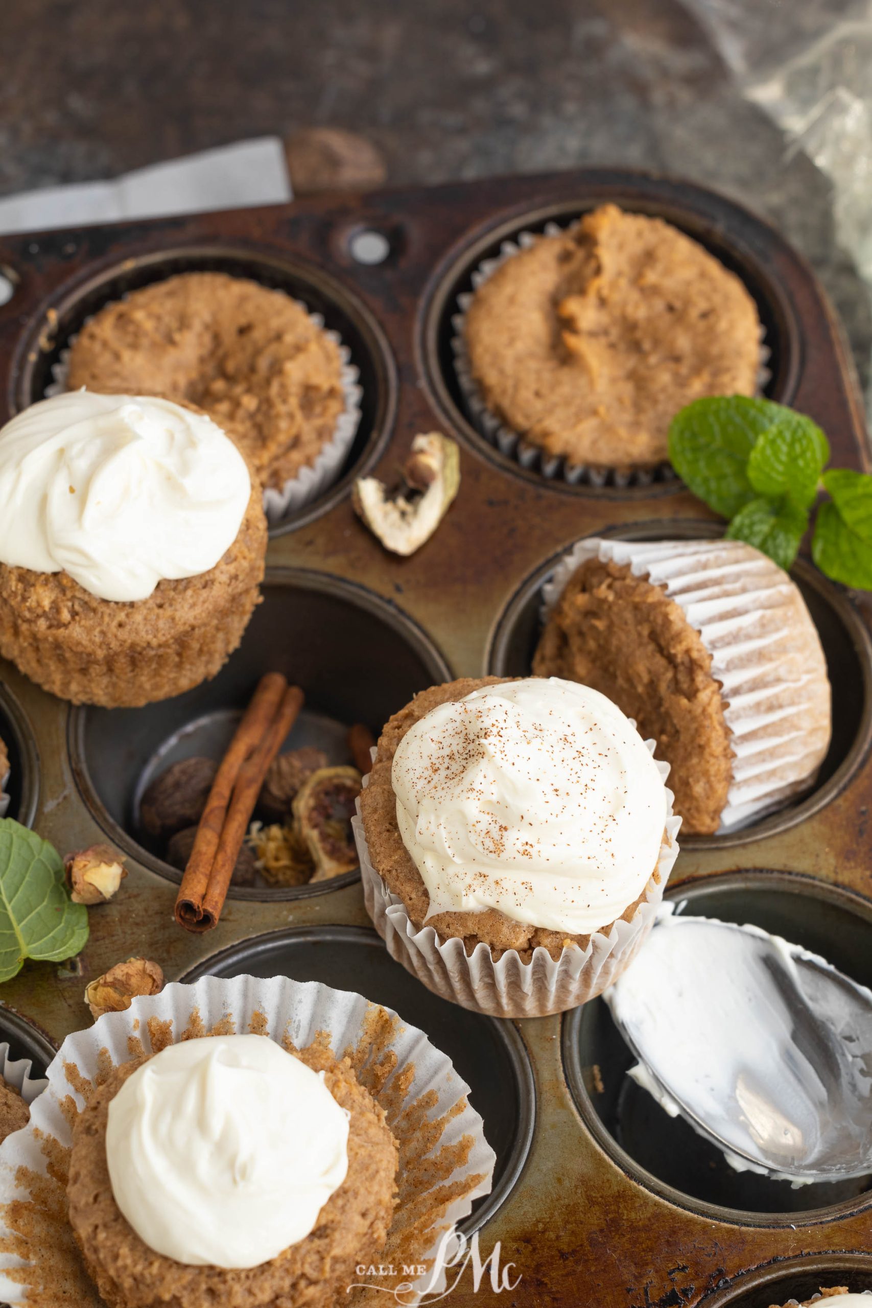 Cupcakes with white frosting in a muffin tray. Some are unfrosted. Cinnamon sticks and green leaves are scattered around.