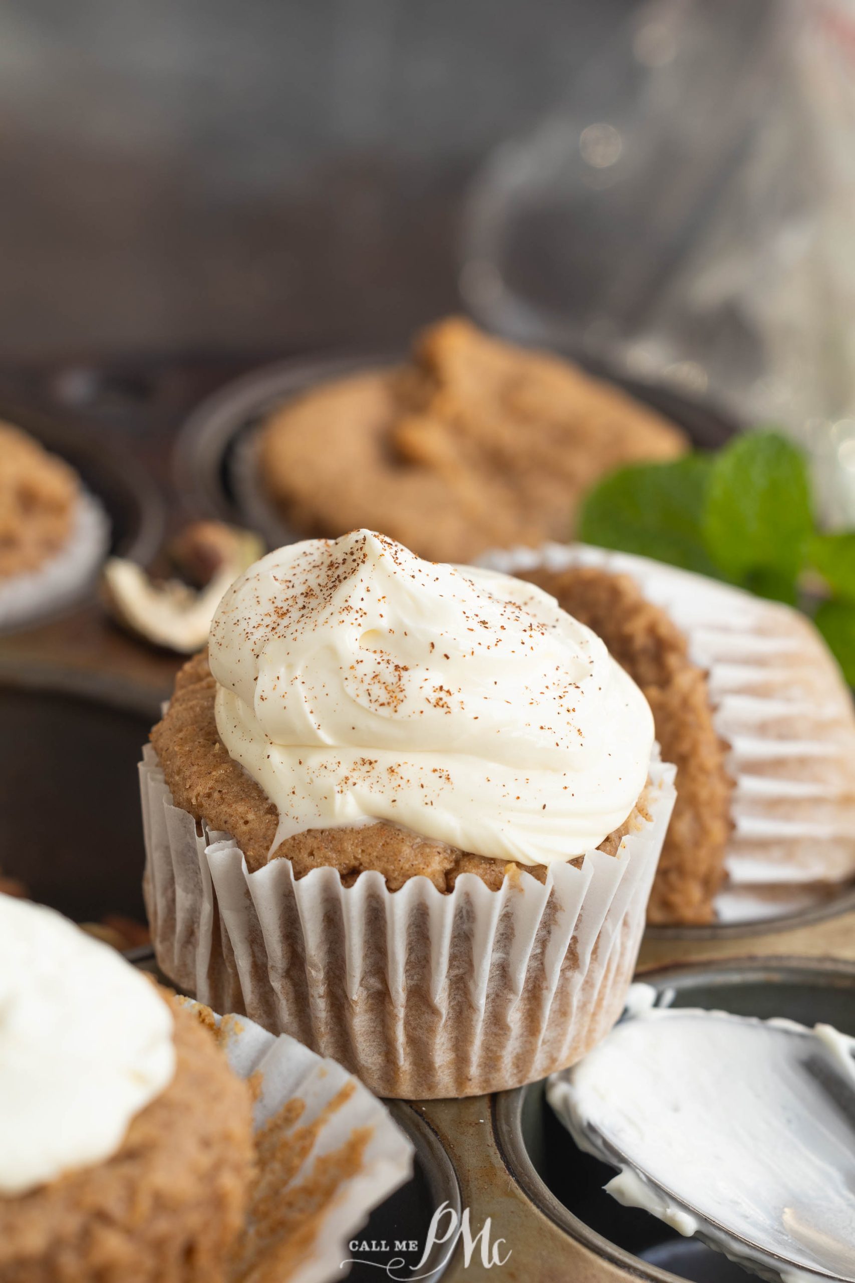 A 2-ingredient spice muffins with white frosting and a sprinkle of cinnamon sits in a muffin tin, surrounded by other unfrosted cupcakes and a mint leaf.