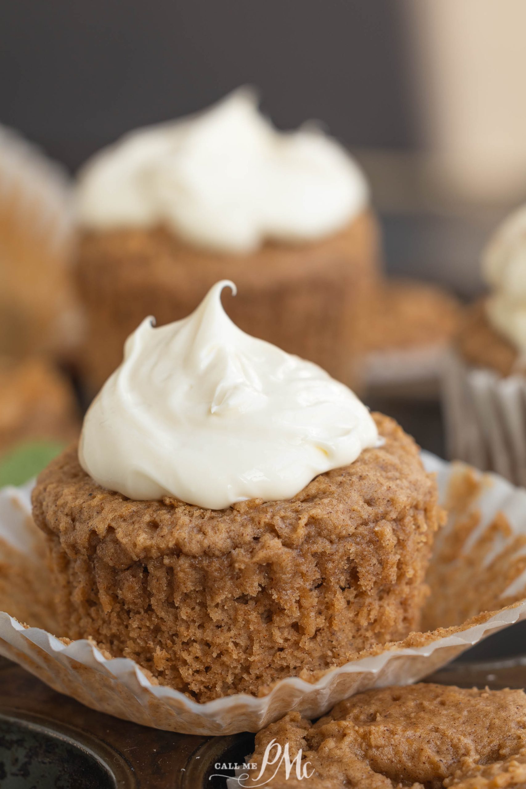 A 2-ingredient spice muffins with white frosting sits in a crinkled wrapper. Other cupcakes are blurred in the background.