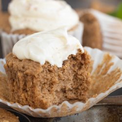 A muffin with cream frosting, partially eaten, sits in an unwrapped paper liner in a muffin tray.