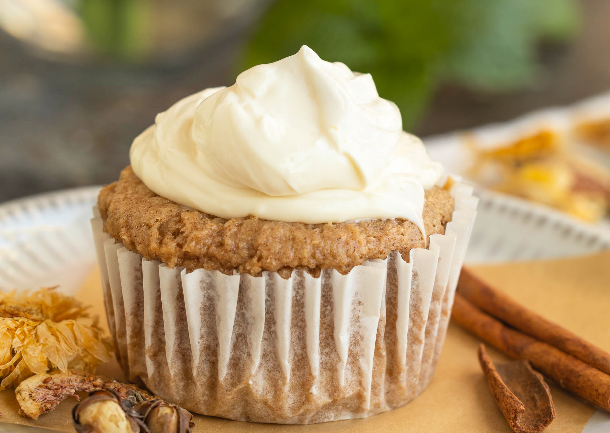 A cupcake with light brown frosting in a white paper liner, garnished with cinnamon sticks and walnuts on a plate.