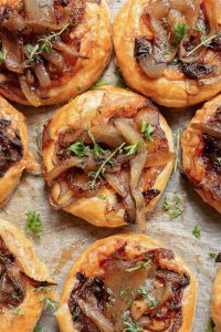 Golden pastry tarts topped with caramelized onions and fresh herbs arranged on parchment paper.