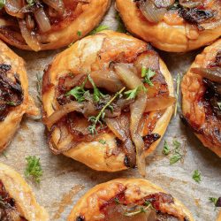 Golden pastry tarts topped with caramelized onions and fresh herbs arranged on parchment paper.