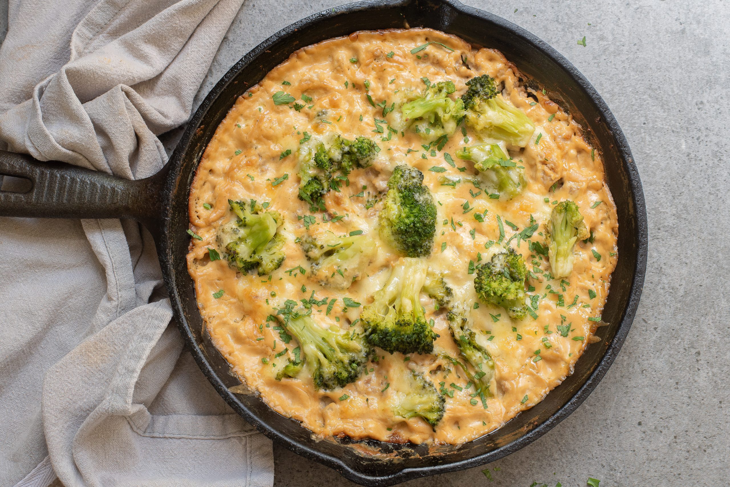 A cast iron skillet filled with a creamy broccoli and cheese dish placed on a gray surface, alongside a gray cloth.