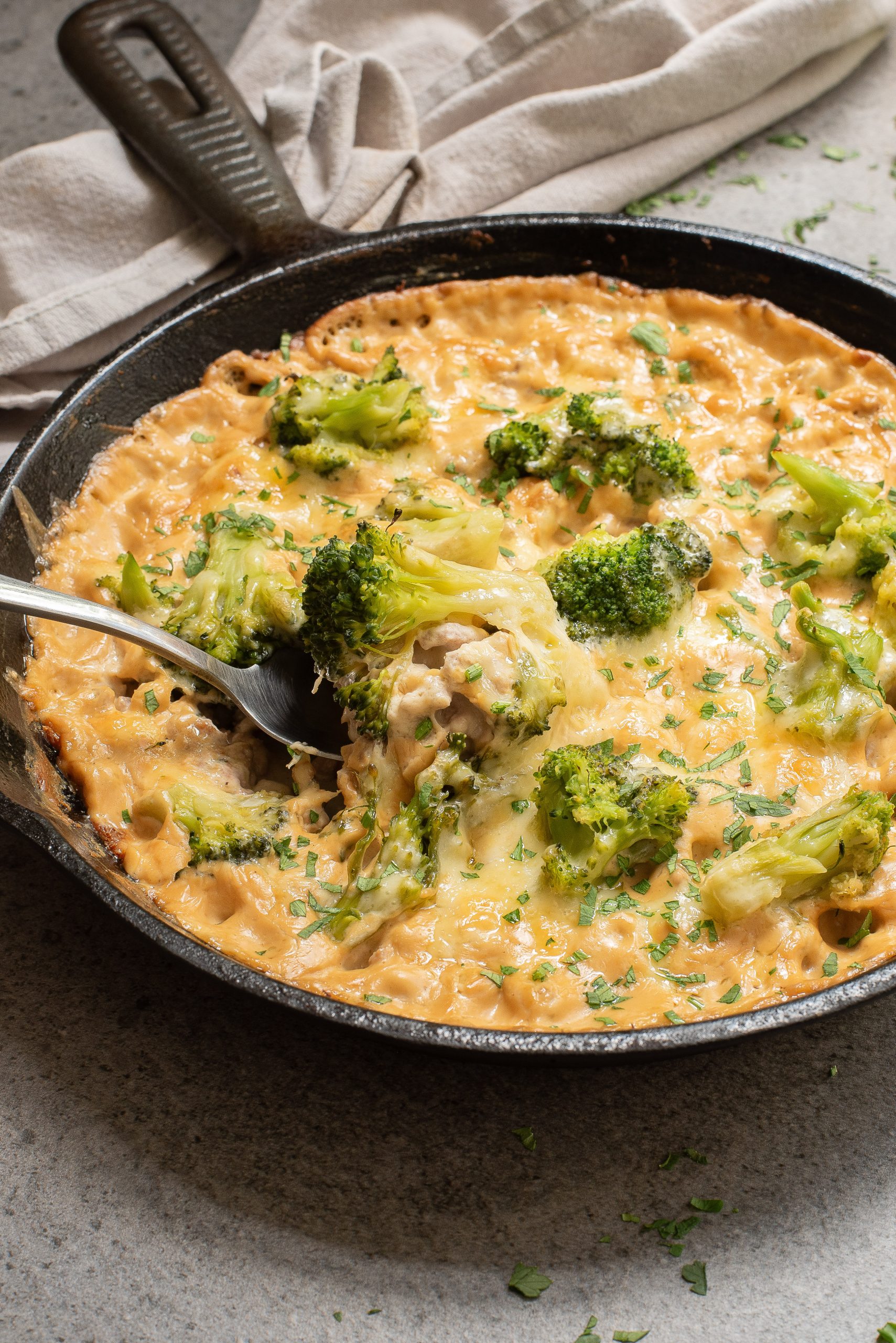 A skillet filled with a creamy casserole featuring melted cheese and broccoli, with a spoon serving a portion. A cloth is in the background on a gray surface.