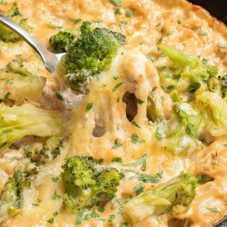 A skillet filled with cheesy broccoli and rice casserole, topped with melted cheese and parsley. A spoon is lifting a portion from the dish.