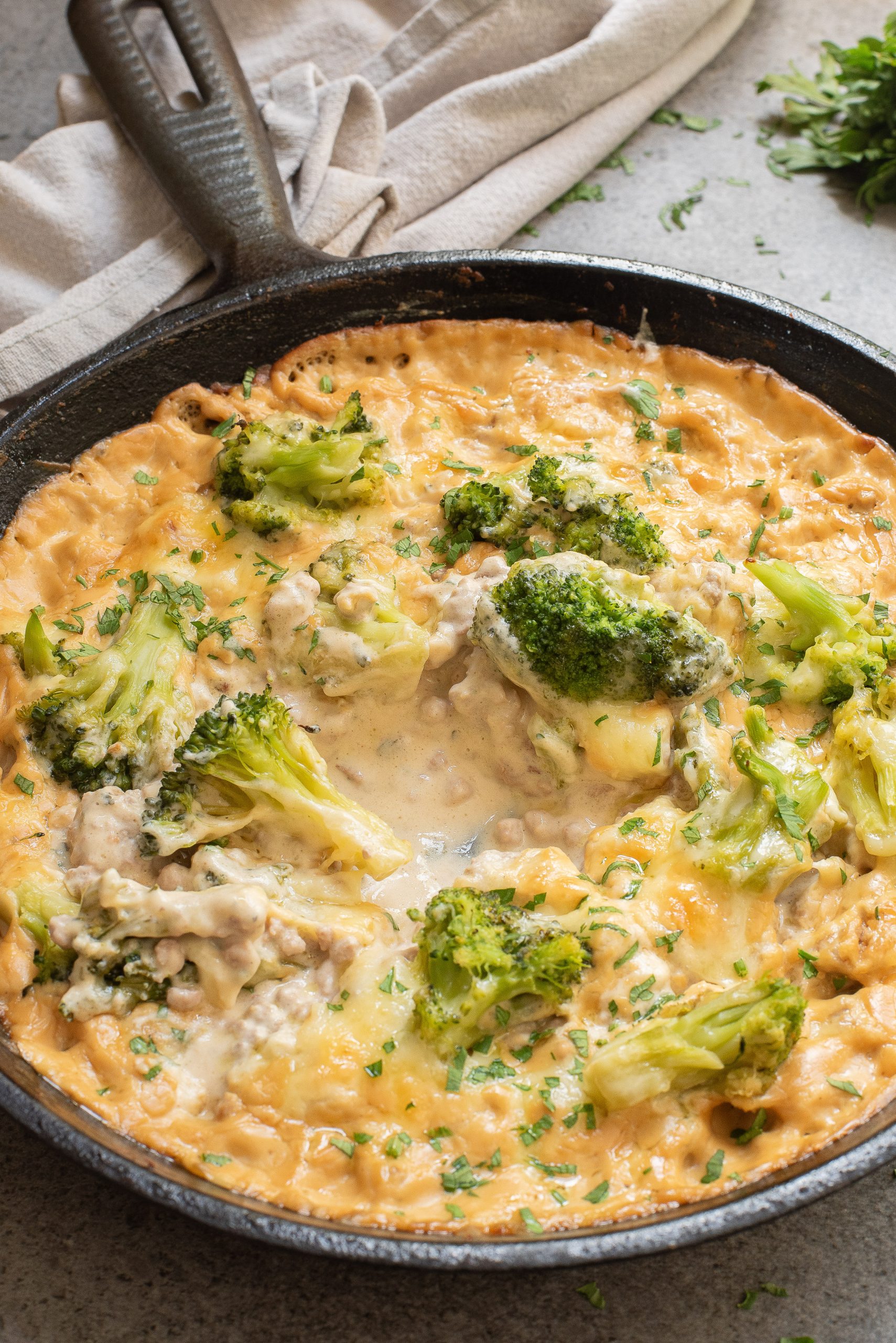 A skillet containing a cheesy broccoli and chicken casserole, garnished with chopped herbs. A beige cloth napkin is placed beside the skillet.