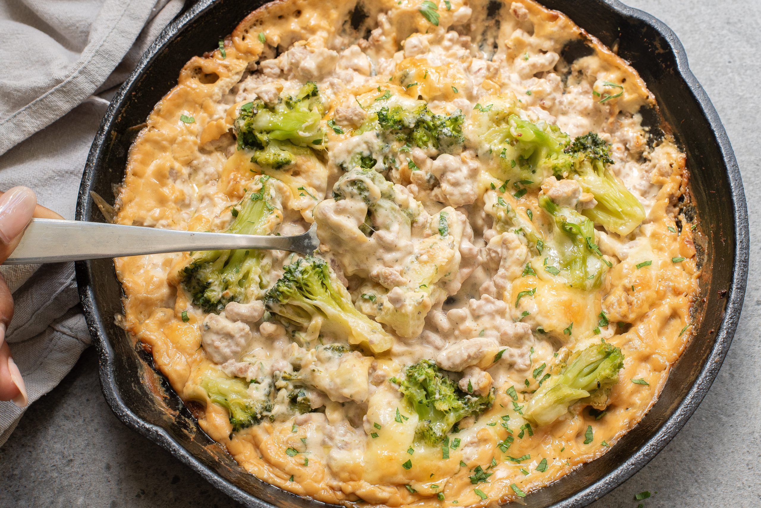 A skillet filled with a creamy, baked broccoli and cheese dish, being served with a spoon, on a gray surface.