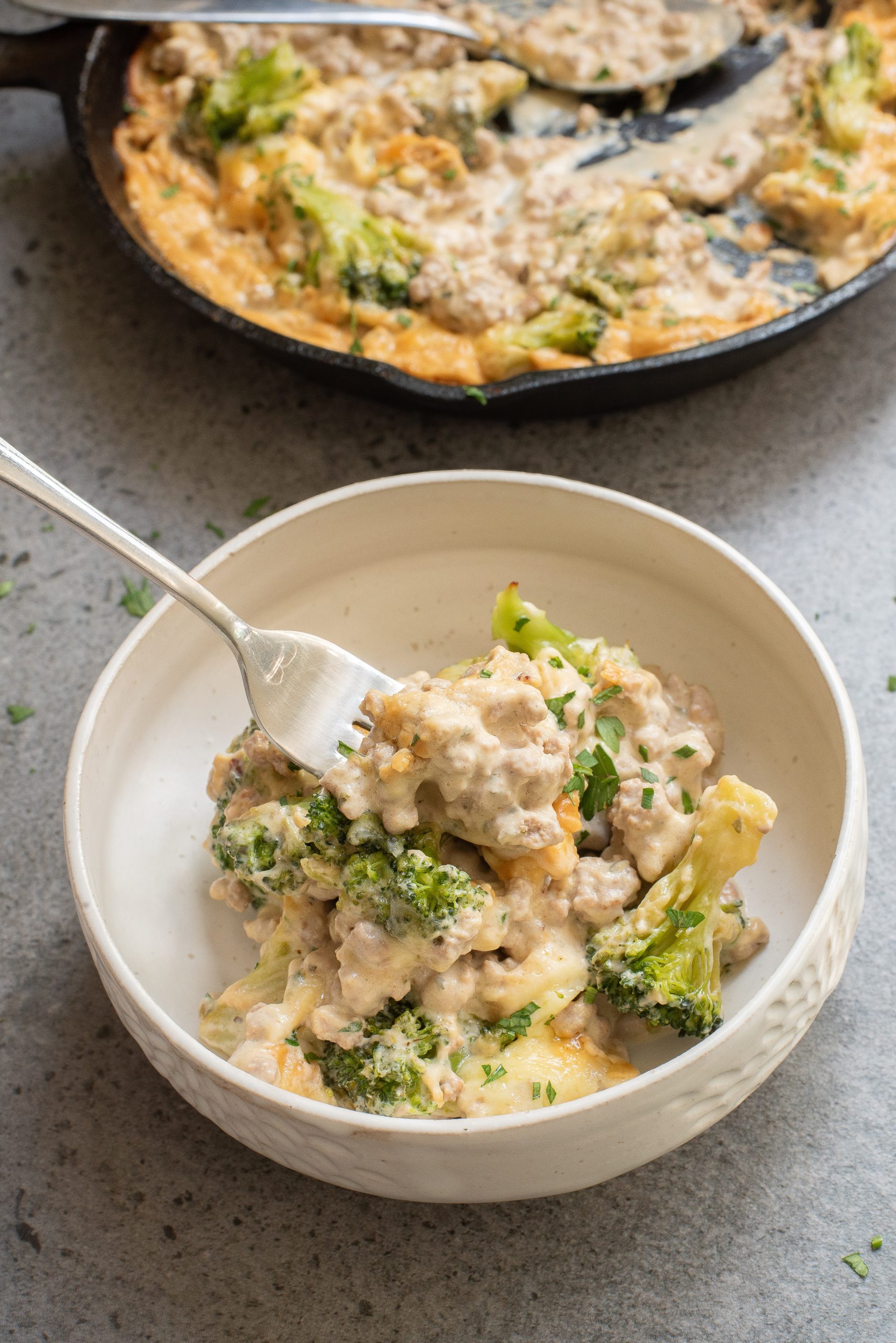 A bowl of cheesy ground beef and broccoli casserole on a table, with a fork resting on top. A pan with more casserole is in the background.