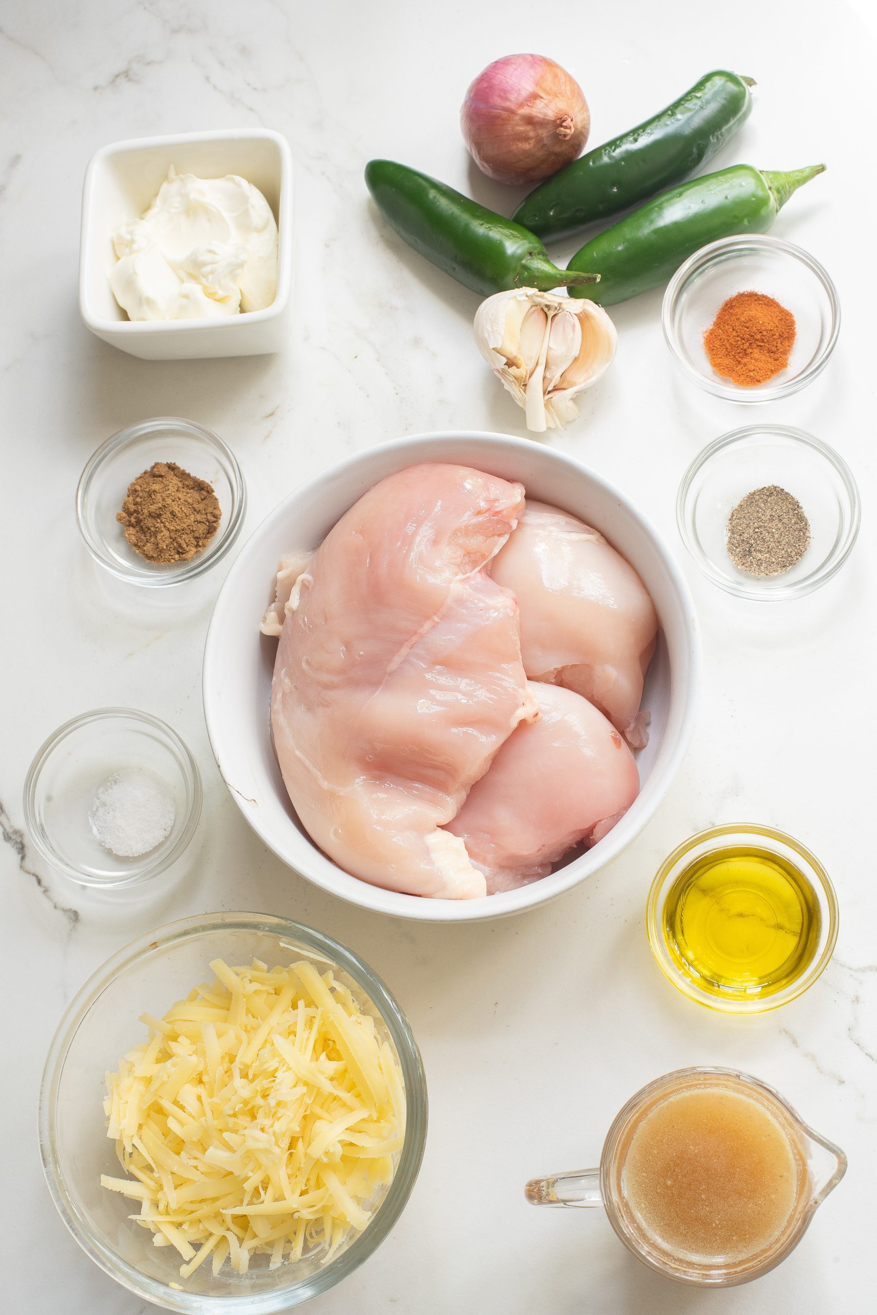 Ingredients on a table: raw chicken breasts, shredded cheese, cream cheese, olive oil, chicken broth, garlic, shallot, jalapeños, and spices.