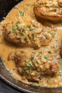 Chicken breasts in a creamy sauce with herbs and sliced vegetables, cooked in a black skillet.