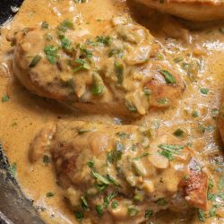 Chicken breasts in a creamy sauce with herbs and sliced vegetables, cooked in a black skillet.