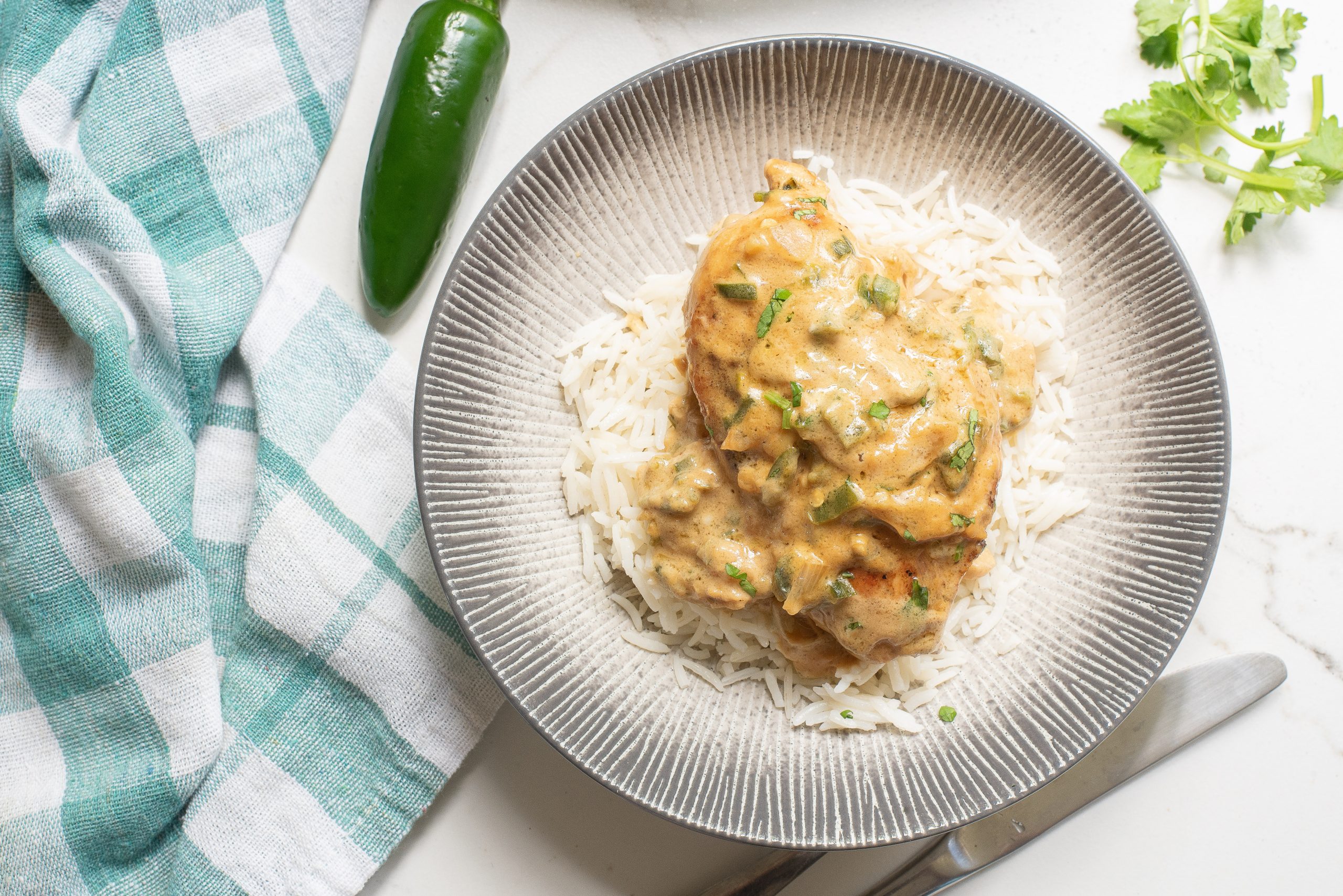 A plate of rice topped with cKeto Jalapeno Cheesy Chicken, garnished with herbs. A green chili, cilantro, and a checkered cloth are nearby.