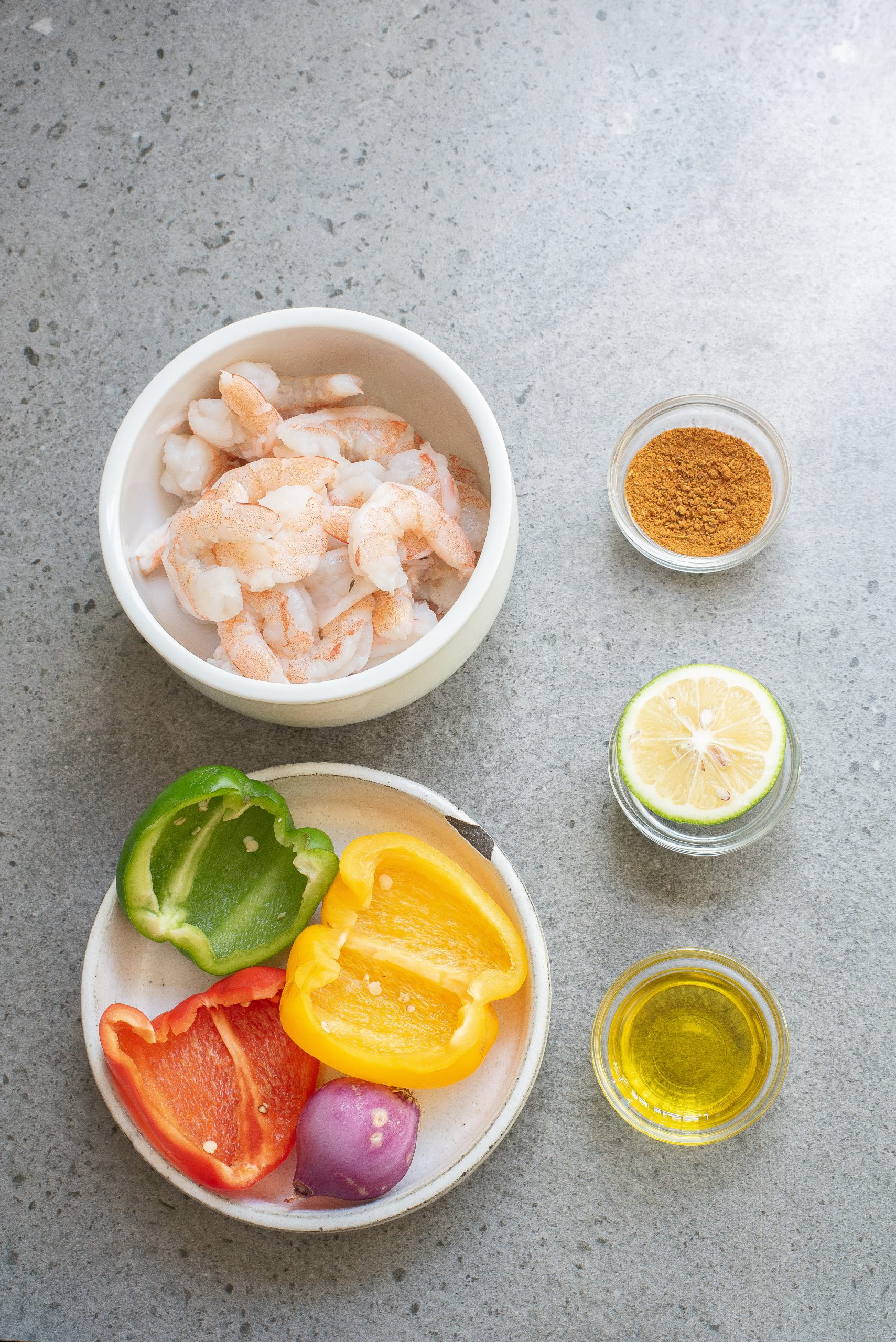 Bowl of shrimp, small bowls of spice and oil, half a lime, and a plate with sliced green, yellow, red peppers, and a small onion on a gray surface.